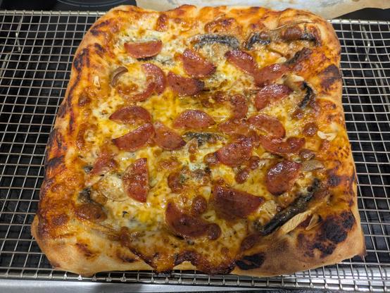 A square shaped pizza sitting on a cooling rack, which itself is sitting on a black cooktop. There are some black spots at the edges, and a small amount of grease in the centre.