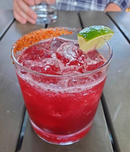 A vibrant red mocktail in a clear glass, filled with ice and topped with a lime wedge. The rim of the glass is coated with chili salt. The drink sits on a dark wooden table at an outdoor setting. In the blurred background, a person's hand holds another glass