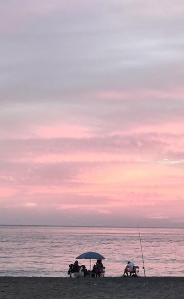 Atardecer en la playa de Salobreña. El sol colorea las nubes de tonos rosas y rojizos. Unas personas están sentadas a la orilla, debajo de una sombrilla.