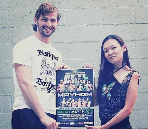 Medium shot of a Caucasian man and an Asian woman standing in front of a textured, light gray brick wall. The man, on the left side of the frame, has light brown hair and a beard, wearing a white t-shirt. The woman, on the right, has long dark hair and is wearing a dark colored pattern top. They both hold a framed promotional poster for "AFC 18 Mayhem." The poster displays images of mixed martial arts fighters and text details about the event. The image has a slightly desaturated, vintage feel. Composition is balanced, with subjects centered in the frame.