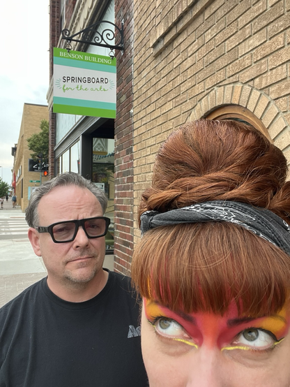While balancing an impressive amount of hair atop her head, an artist snaps a bad selfie to document her suave photographer husband underneath the Springboard for the Arts exterior sign.