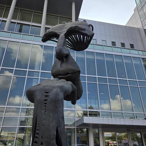 Photograph of a statue that has a stylized hand leaning on a chin. However, the entire face is not seen in the status - only the chin, lips, nose, and eye socket. The rest of the head is not on the statue. The statue is in front of a tall building with many, many windows.
