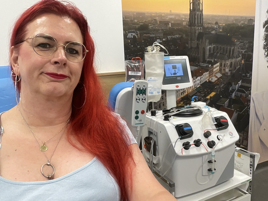 Emma, met rood haar en bril, glimlacht ontspannen tijdens haar plasmadonatie bij de bloedbank. Achter haar zijn medische apparaten te zien die gebruikt worden voor het afnemen van plasma. Ze draagt een mintgroene top met uitgesneden hals.