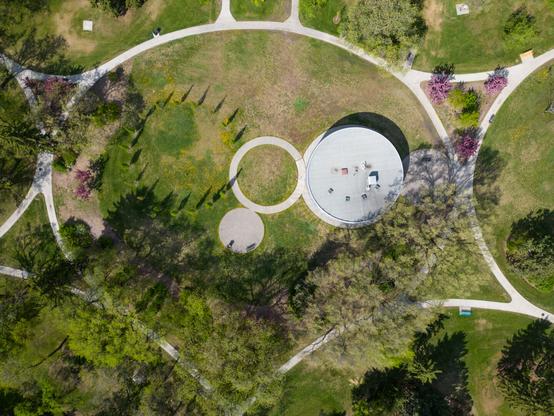 Aerial view of Borden Park in Edmonton during spring. The photo shows a circular pavilion structure with a flat white roof surrounded by winding walking paths and green lawns. Trees in various stages of bloom cast long shadows across the park, and the paths form clean geometric shapes that contrast with the organic patterns of nature.