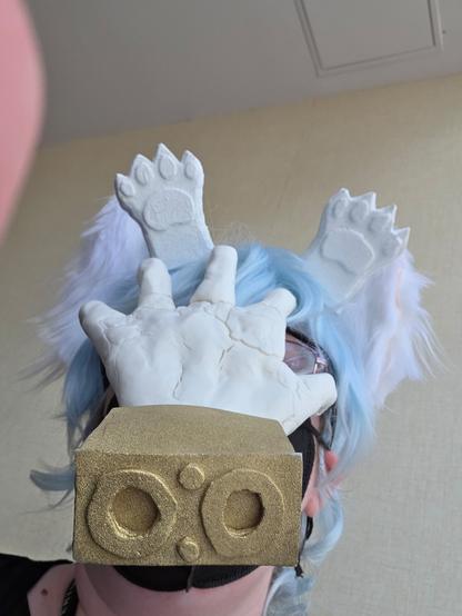 Closeup photo of my head with the face covered by a white disembodied hand with a gold wrist part, based on the one Tomura Shigaraki wears in My Hero Academia. I am also wearing a short light blue wig, fluffy white cat ears, and a pair of disembodied white paws in front of the cat ears. Both the paws and the hand part have cracked surface texture.