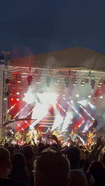 A live music performance on stage with bright lights and smoke effects. Several musicians are visible, and a large crowd is enjoying the show. The atmosphere is vibrant and energetic, set against a night sky.