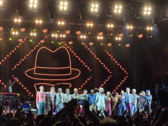 A large group of musicians and performers stands on stage, smiling and posing together, with bright stage lights illuminating the scene. A large, stylized graphic of a hat is displayed in the background, while audience members capture the moment with their phones.