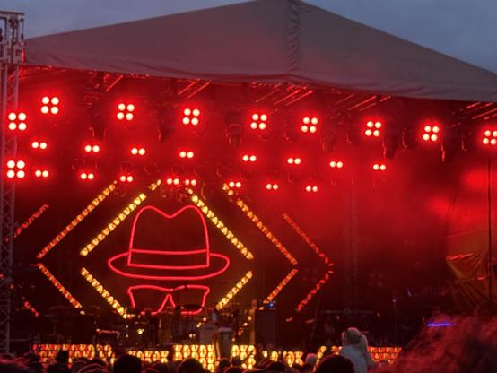The image shows a stage at a concert with dramatic red lighting. A large neon outline of a hat and glasses is displayed in the background, with various instruments and equipment visible in the foreground. The crowd is partially visible in front of the stage.