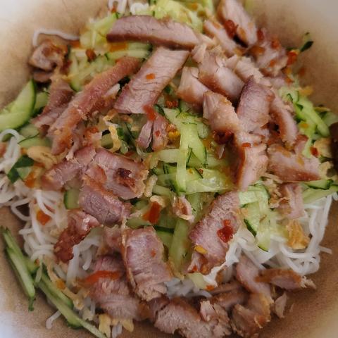 Top-down close-up of a rice noodle salad in a bowl. The salad consists of thin, white rice noodles topped with sliced grilled pork, julienned cucumber, and small pieces of crispy fried shallots. Red chili flakes are scattered throughout.