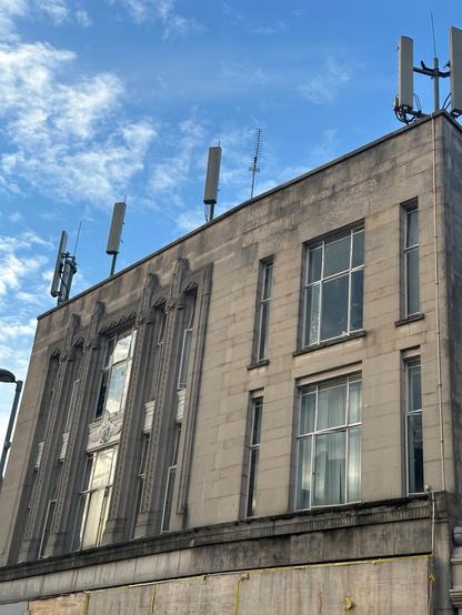 the two upper stories on a stone-clad interwar modernist building in London, showing columns of tall, slender vertical windows in an art deco idiom. The stone is grimy in places. An array of mobile phone base station aerials lines the roof.