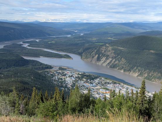 A view from the top of a mountain looking down at the wide and silty Yukon River, with Dawson City on its banks below. Another river - the Klondike - with clear water is flowing into it, creating a dark area where it merges with the muddier and larger Yukon River.