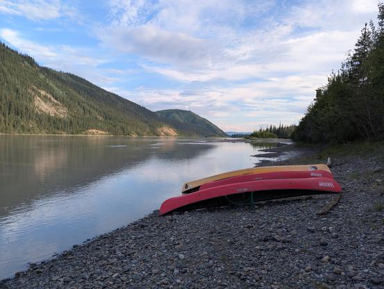A photo of three canoes (two red, one yellow) on the rocky shore of a wide river. The mountains in the background and cloudy sky are reflected in the river. The sun is low but still bright.