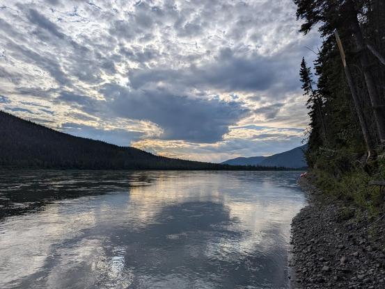 A photo of a cloudy sky with the sun behind, reflected on a wide river lines with mountains and forest.