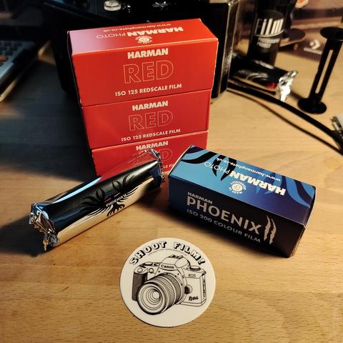 3 boxes of 120 Harman RED, 1 box of 120 Harman Phoenix II, and a roll of 120 Harmon Phoenix II out of the box but still in it's silver packaging.
Near the bottom of the image is a sticker that reads "SHOOT FILM!" above an illustration of a Canon EOS Rebel.