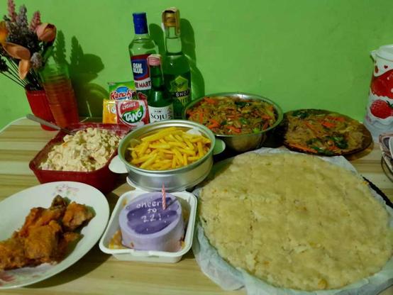 Table with various food. From top left, clockwise: Pinoy fruit salad, french fries, two bowls of pancit bihon noodles, maja blanca pudding, small chocolate cake, fried chicken. At the vaxk is a vase of faux flowers and bottles of alcoholic drinks