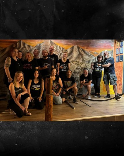 Gruppenbild von Metalheads in einer urigen Kneipe. Sie stehen oder hocken vor einem Wandbild, das Gebirgsgipfel zeigt.