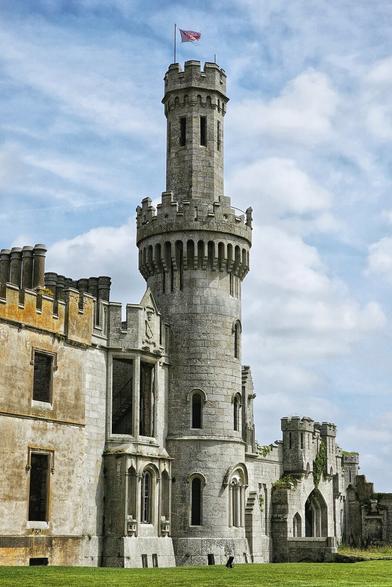A magnificent, imposing view of Duckett's Grove, a Gothic Revival mansion in County Carlow, Ireland, stands under a bright, cloudy sky. The main focus is the elaborate central tower, constructed from light-coloured stone, with intricate detailing, arched windows, and a crenellated top. To the left and right, the substantial remains of the mansion's walls, with their distinctive pointed arches and decorative stonework, convey a sense of its former grandeur despite their ruinous state. Lush green grass stretches across the foreground, leading up to the historic structure, which evokes a strong sense of Irish heritage and romantic decay.
