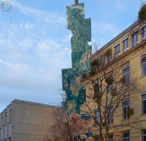 Vertikales an Mast im Straßenraum der Wiener Franklinstraße befestigtes hohes Kunstwerk aus versetzt angeordneten Rechtecken, das einen Flussverlauf auf Luftbild zeigt