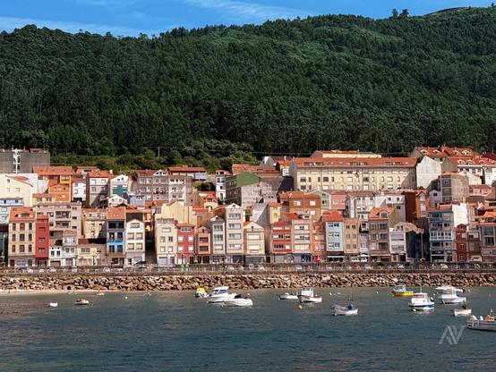 La imagen muestra una villa costera con casas coloridas alineadas frente al mar. Las viviendas están pintadas en tonos variados como rojo, azul, amarillo, naranja y blanco, y tienen tejados de tejas rojas. Delante de ellas hay un rompeolas de piedras grandes y un grupo de pequeñas embarcaciones blancas flotando en el agua. Al fondo, se eleva una colina densamente cubierta de árboles verdes. El cielo es azul claro.