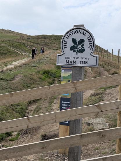 Sign for Mam Tor behind wooden fence 
People walking on stone path up hillside