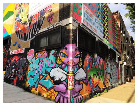 2019 photo of street art on a building in Queens, NY by Shiro (lower level wraparound), ELLE (upper left), and Garrison Buxton (upper right) — all part of the Welling Court Mural Project, a community beautification initiative. (Buxton’s art also extends past left frame and features rainbow-colored stripes for Pride.) Find out more about the artists on their Instagram accounts: @shiro_one, @ellestreetart, and @thegarrisonbuxton.

Image description: A building exterior covered in street art, with the corner at the center of the photo and two walls receding towards left and right. On the first floor, wrapping around the corner to both sides, is a purple-skinned female figure with exaggerated cherubic features and makeup. Her open palms are spread toward the viewer. Hovering between them, like an offering, is a spray can with white feathered wings, labeled with the text LOVE IS ALL (with “is” in white letters inside a red heart). To her left is the artist’s tag SHIRO in giant stylized letters; to her right is the giant word YES! Both words are done in a variety of vibrant colors. Additional artwork is visible on the walls as they recede. On the second floor at left is a circular image of abstract shapes and colors, partially hidden by a commercial sign. At upper right is a grid of diamonds in rainbow colors, partly obscured by a sign for ARTAMUS RESTAURANT EQUIPMENT & SUPPLIES.