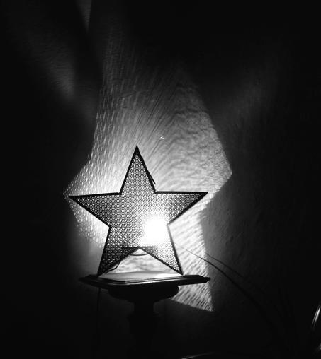 A star-shaped black metal lamp casts geometric shadows onto the walls of a dark room. (Version françæ) Æn lampe en métal noirx en forme d’étoile projette des ombres géométriques sur læ mur dal pièce plongeæ dans l’ombre.