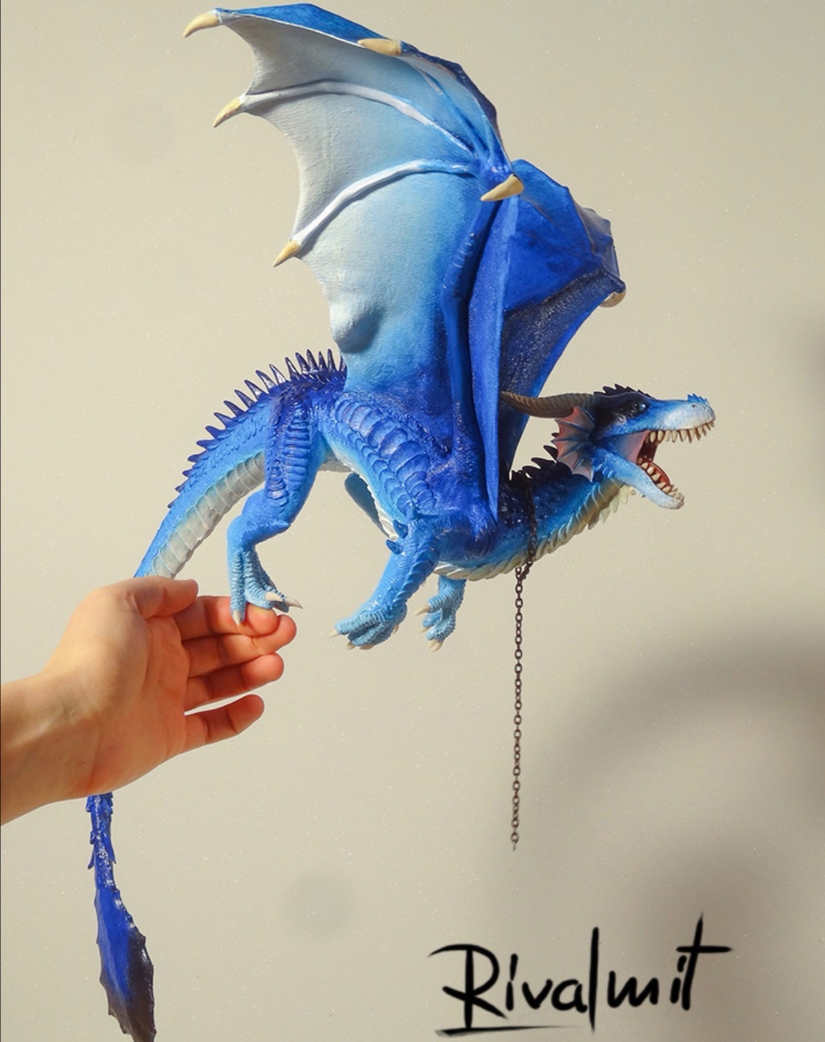 Photo of a Sculpture of a big blue dragon.  It's freely balancing on the hand which is juuust on the side of the picture. the pose is wings open, bowed forwards as if its ready to jump.