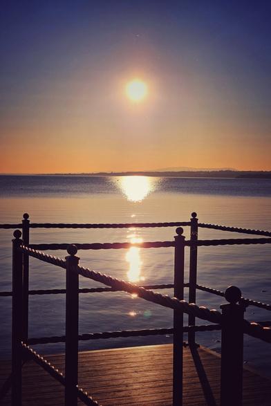 A serene lakeside view at sunset, looking out from a wooden pier with a rope railing. The sun, a bright orb, is setting over the distant horizon, casting a shimmering golden reflection across the calm water. The sky transitions from a deep blue at the top to a warm orange and yellow near the horizon. Silhouetted hills are faintly visible in the distance.