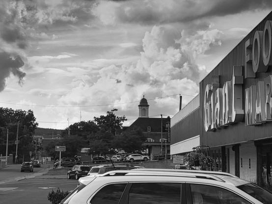 A black and white street scene captures a small town’s main thoroughfare under a dramatic sky of billowing cumulus clouds. The wide street stretches into the distance, flanked by parked cars along both sides and marked by painted lane dividers down the center. On the right, a large commercial building dominates the frame with bold lettering spelling “FOOD MART” across its corrugated metal facade, while smaller storefronts with awnings extend beyond it. A church steeple with a distinctive cupola rises in the middle distance, its dark silhouette contrasting against the bright sky. Mature trees line the street, their full canopies creating pools of shadow that dapple the pavement. Power lines stretch across the frame, adding geometric elements to the organic shapes of clouds and foliage. The lighting appears natural and diffused, creating strong tonal contrasts between the bright sky, shadowed storefronts, and sun-dappled asphalt that give the scene depth and visual weight.