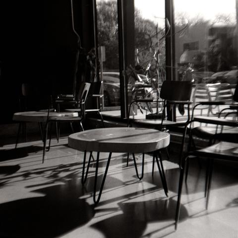 Black and white photograph of bright light streaming through tall windows casting long shadows from small wooden tables and chairs on a concrete floor.