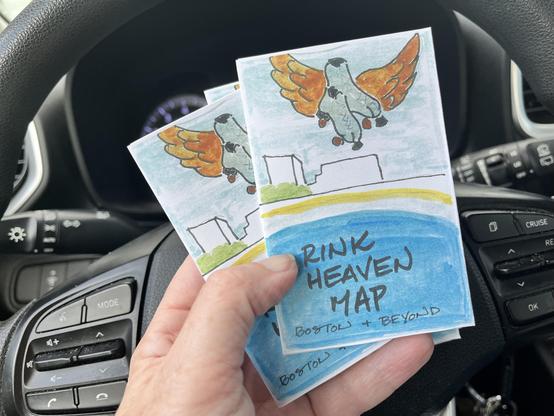 a hand holds a number of small, eight fold zines. The zine cover reads "Rink Heaven Map" and shows a watercolor of a rink with some winged skates floating in the sky. In the background you see a car steering wheel and dashboard; the person holding the zines is apparently in the drivers seat of a parked car.