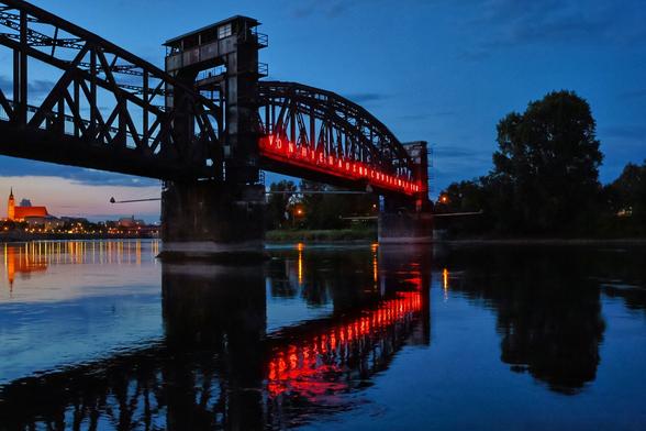 A nighttime long shot captures the Hubbrücke bridge over the Elbe river in Magdeburg, Germany. The dark, sturdy iron railway bridge spans the serene water, which perfectly mirrors the vibrant red illuminated text "Vᴏɴ ʜɪᴇʀ ᴀᴜs ɴᴏᴄʜ ᴠɪᴇʟ ᴡᴇɪᴛᴇʀ" (FROM HERE MUCH FURTHER) displayed along the bridge's arch. On the left bank, the distant lights of Magdeburg's city skyline and the distinctive towers of the Magdeburg Cathedral are subtly visible under a dark blue twilight sky. The scene evokes a sense of calm and reflection, highlighting the architectural presence and its illuminated art installation.