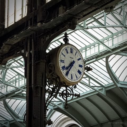 Une vieille horloge de la Gare de Lyon accroché aux poutres en métal. Dans le fond, une verrière est éclairée.