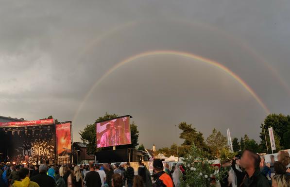 OpenAir Konzert Noisehausen in Schrobenhausen. Links befindet sich eine Bühne auf dem die Band "Das Lumpenpack" spielt. Rechts davon befindet sich ein doppelter Regenbogen am Himmel.