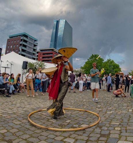 in einem Holz steht eine asiatisch anmutende Person mit einem Strohhut auf dem Kopf und zweien in der Hand. Sie jongliert damit. Im Hintergrund jongliert ein Mann in kurzen weißen Hosen mit kegeln.