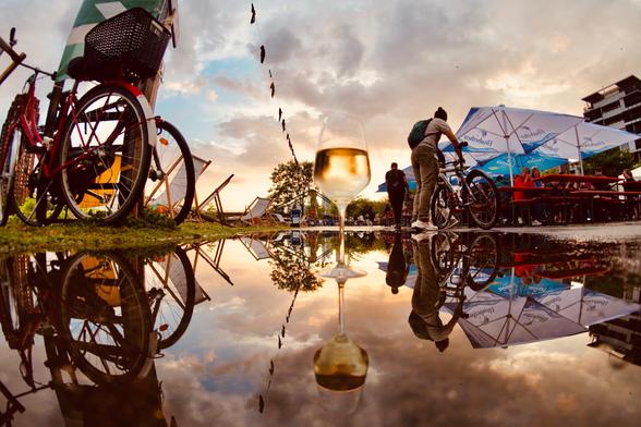 eine Spiegelung in einer Pfütze. Links ein Fahrrad hinten Sonnenuntergang, Himmel mit Wolken und in der Mitte ein Weinglas mitten in der Pfütze.