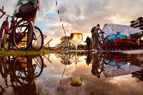 eine Spiegelung in einer Pfütze. Links ein Fahrrad hinten Sonnenuntergang, Himmel mit Wolken und in der Mitte ein Weinglas mitten in der Pfütze.