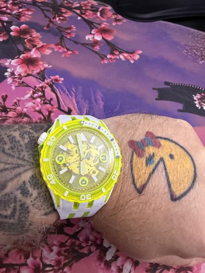 The image features a close-up of a person's wrist wearing a bright yellow and white wristwatch with a transparent face, showcasing the inner workings of the watch. The watch has a yellow bezel with white markings and a white strap with green accents. The person's wrist also displays a tattoo of a yellow character with a red bow on its head, resembling a cartoonish animal. The background includes a fabric with a floral pattern, predominantly pink cherry blossoms against a purple sky, and a silhouette of a traditional building. The overall composition highlights the vibrant colors of the watch and the tattoo against the floral backdrop.