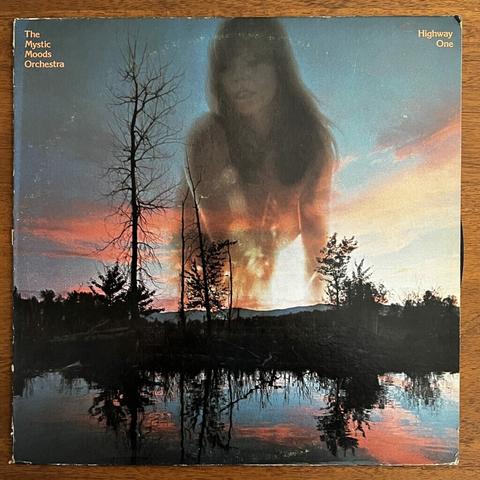 Mystic Moods Orchestra front cover – with a dynamically colored photograph of a reflective lake at dusk, surrounded by some wooded hills, trees both leafed and bare, and, superimposed on a red sunset sky, a photograph of a lovely naked woman with long flowing straight brown hair and bangs.