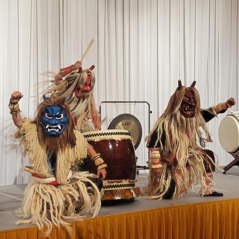 Three performers in elaborate "Namahage" demon costumes play traditional Japanese taiko drums on a stage. Each performer wears a fierce-looking demon mask—blue, red, and brown—with long hair and straw garments. Two are kneeling and raising clenched fists, while the third stands behind them, mid-drumbeat. The backdrop is a white curtain and the stage is decorated with large Japanese drums and a gong.