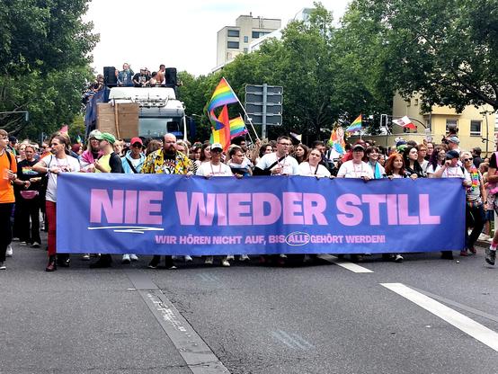Das Bild zeigt eine Gruppe von Menschen, die an einem Umzug oder einer Demonstration teilnehmen. Sie tragen ein großes Banner mit der Aufschrift „NIE WIEDER STILL. WIR HÖREN NICHT AUF, BIS ALLE GEHÖRT WERDEN!“.  Der Slogan bedeutet, dass sie nicht aufhören werden, bis alle gehört werden.  Es ist ein starkes Statement für Gleichberechtigung und gegen Diskriminierung, wahrscheinlich im Kontext einer LGBTQ+-Demonstration.  Im Hintergrund sind Regenbogenfahnen zu sehen, was die Vermutung unterstützt. Viele Teilnehmer tragen Regenbogen-Accessoires oder Kleidung. Die Atmosphäre wirkt engagiert und positiv.