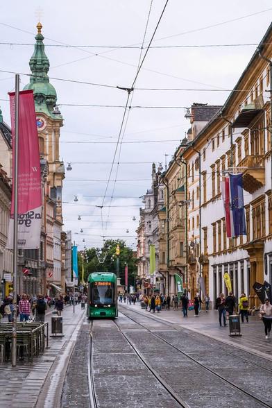 Hochformatiges Foto der Grazer Herrengasse. In der Mitte sind Straßenbahn-Gleise, es kommt eine Straßenbahn entgegen. Rechts und links befinden sich Fußgängerzonen mit vielen Menschen.