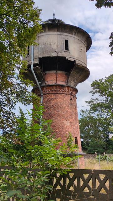 A ruined water tower. It consists of a narrowing column made of red brick, on top of which a cylindrical, gray concrete tank has been built, with its bottom supported on an arched concrete. The front sections of this arched support fell off, revealing the hidden top of column, and the darkness inside the tank. To the left, another part of the support seems to be falling apart, supported only on gutter pipe that's mounted to the concrete just above the rift, and goes from top of the tower to its bottom. Its very top seems to be finished with something that resembles an umbrella, with a spike on top. At the bottom of the column there's an arched window, and in the tank there are two narrow rectangular windows that aren't particularly pretty. Around it, trees. The tower is surrounded by a mesh fence, while the whole plot overgrown with grass is surrounded by a concrete fence with X-shaped pattern.