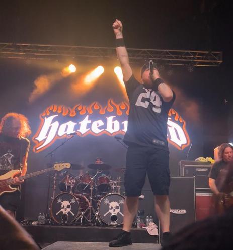 A photo of Jamey Jasta from Hatebreed on stage holding a mic with his fist twisted in the air performing live. A banner behind him reads Hatebreed. There’s a drum kit behind him and other band members beside him. Stage lights shining bright around them all.