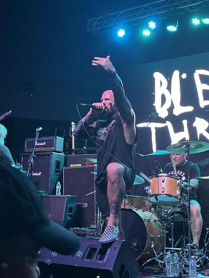 A photo of Brandan from Bleeding Through performing live holding a microphone. Blue and green stage lights shining above him. A banner behind him reads Bleeding Through.