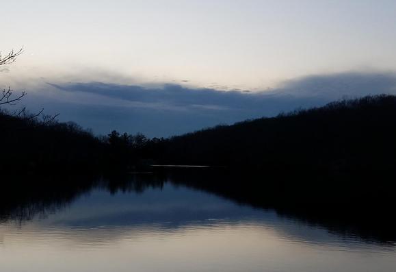 A soft gray twilight long past sunset. Standing beside a pond, looking across its lightly rippled waters. The far shore is wooded, the forest silhouetted in the fading light. Although no snow or ice is visible, it is the heart of winter. The tree branches are bare except for a few evergreens. A thick bank of clouds along the horizon is shades of darker gray against the clear skies above. The sky and silhouetted forest are mirrored on the pond below.