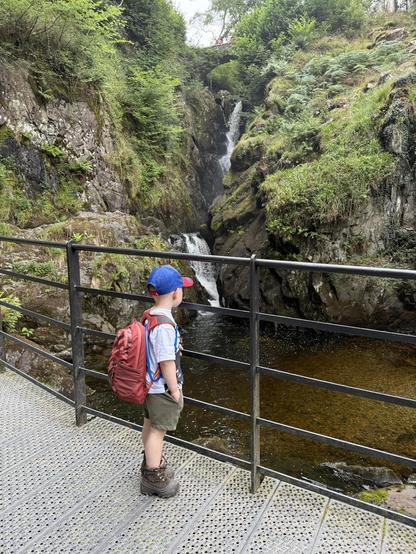 A young boy stands on a metal walkway near a cascading waterfall, surrounded by lush green vegetation and rocky cliffs. He wears a red backpack, a blue cap, and brown hiking boots. The scene captures a serene natural setting.