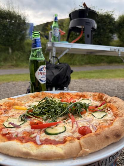 A freshly made pizza topped with cheese, sliced vegetables, and rocket sits in the foreground, with two bottles of green beer behind it. A portable grill is visible on a table, set against a backdrop of greenery and cloudy skies.