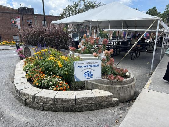 Color photo of plantings, public seating, and shade tents in a public right of way. A prominent sign boasts of the efforts to make the community more “ADA ACCESSIBLE”