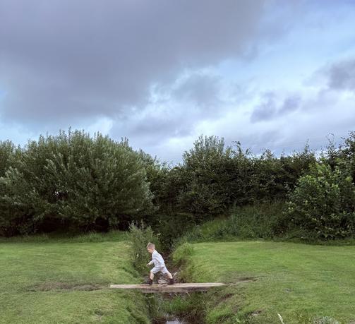 A child in light clothing is walking across a wooden plank bridge over a small stream in a grassy area. The scene is surrounded by trees and cloudy skies.